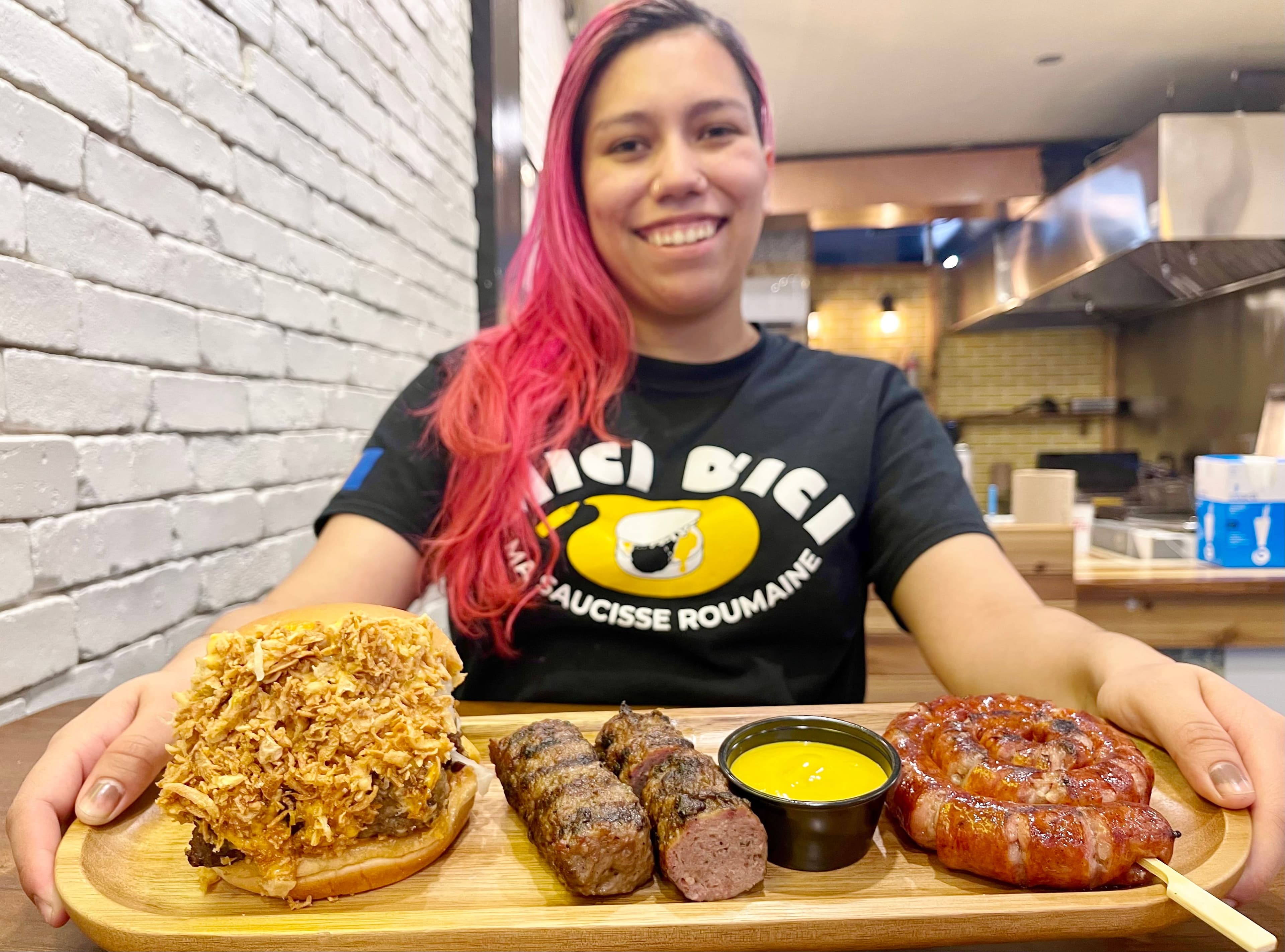 Une employée de Mici d’ici : ma saucisse roumaine, Lina Piquette, tient un plateau contenant un burger double Cannon comportant deux mici et de l’oignon frit, deux mici au centre, et à droite un sausuçon.